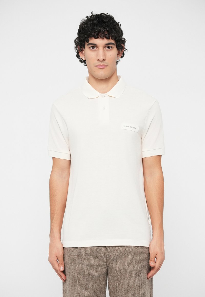 Νεαρός άντρας με σγουρά μαλλιά, φορώντας ένα απλό λευκό polo shirt μ ε ετικέτα "Armani Exchange" και ανοιχτόχρωμο καφέ υφασμάτινο παντελόνι.