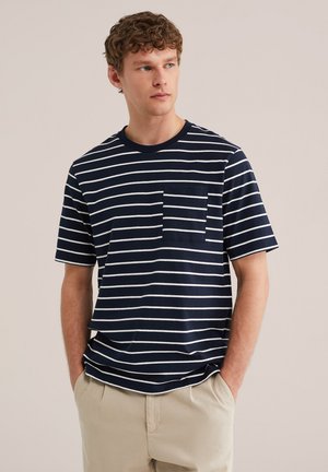 Mlajši moški z kodrastimi lasmi, oblečen v mornarsko modro in belo majico s horizontalnimi črtami, z žepom na prsih in bež hlačami, roke v žepih, gleda v desno.