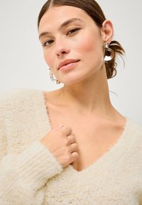 Maglione fluffy avorio con scollatura a V, caratterizzato da polsini a coste. Il modello indossa orecchini a cerchio metallici e anelli d'argento, mostrando una texture liscia.