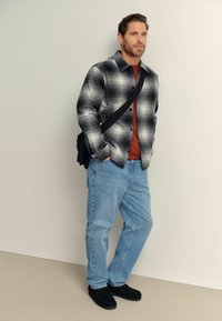 Camicia a quadri bianca e nera, t-shirt rossa sotto, jeans azzurri chiari, scarpe nere e una borsa nera a tracolla. Texture liscia, design casual.