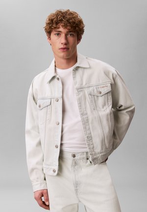 Giovane uomo con capelli ricci che indossa una giacca di jeans bianca, una maglietta bianca e pantaloni bianchi, in piedi davanti a uno sfondo grigio uniforme.