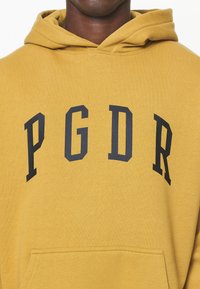 Gelber Kapuzenpullover aus Baumwolle mit einer Fronttasche und fetten marineblauen Buchstaben "PGDR" auf der Brust. Verfügt über eine Kapuze mit Kordelzug und eine weiche Textur.