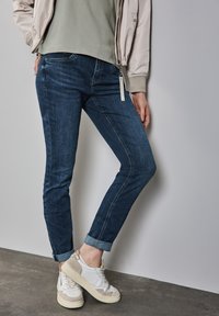 Jean en denim bleu foncé avec une coupe slim et des poignets roulés, associés à des baskets blanches et gris clair. La tenue comprend un haut vert clair et une veste bomber pâle.