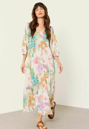 Frau geht drinnen und trägt ein langes, weißes Blumenkleid mit dreiviertel Ärmeln und braunen Sandalen.