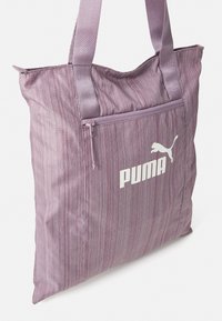 Lila raitapussukka, joka on valmistettu kankaasta, ja siinä on tukevat kahvat, edessä vetoketjullinen tasku sekä valkoinen PUMA-logo sivussa.
