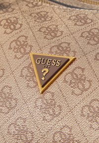 Smešano smeđe tkanje sa teksturiranom završnom obradom sadrži trokutasti smeđi i zlatni akcent logotipa "GUESS" sa upitnikom.