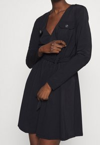 Robe noire à manches longues, décolleté en V, deux poches avant et taille à cordon pour un ajustement modulable, fabriquée dans un tissu léger.