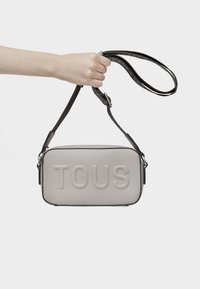 Borsa a tracolla grigia con texture e logo "TOUS" in rilievo, chiusura zip e tracolla nera regolabile. Mano che tiene la tracolla per la visualizzazione.
