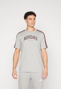 Pelēka kokvilnas T-krekla ar īsām piedurknēm, kuram ir tumši bordo "ADIDAS" logo un horizontālas svītras uz pleciem.