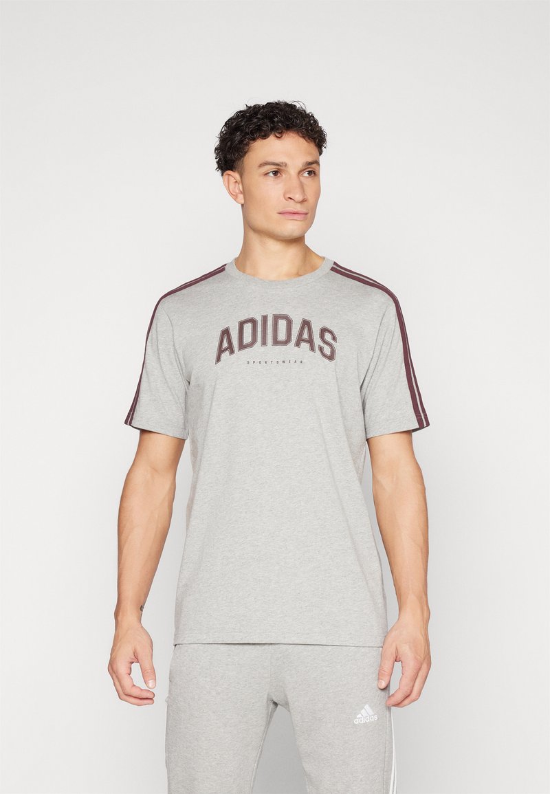 Pelēka kokvilnas T-krekla ar īsām piedurknēm, kuram ir tumši bordo "ADIDAS" logo un horizontālas svītras uz pleciem.