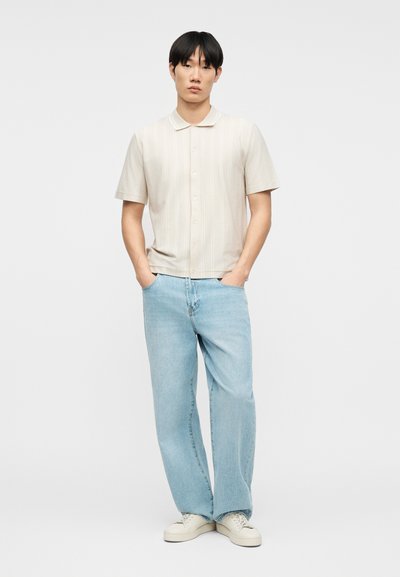 Νέος άνδρας στέκεται με τα χέρια στις τσέπες, φορώντας ένα μπεζ πουκάμισο με κοντά μανίκια και κουμπιά, ανοιχτόχρωμα μπλε τζιν και λευκά αθλητικά παπούτσια, μπροστά σε ένα απλό φόντο.