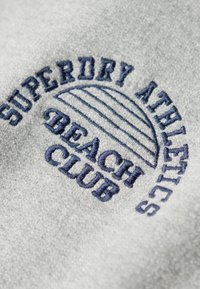 Tela gris con bordado azul oscuro. El texto dice "SUPERDRY ATHLETICS BEACH CLUB" con líneas horizontales y acentos redondeados.