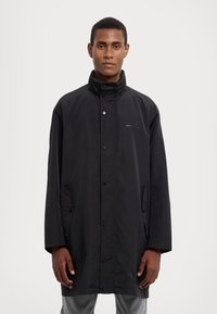 Černý nepromokavý kabát s vysokým límcem, patentkami a bočními kapsami; má hladkou texturu a minimalistický design.