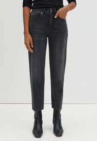 Woman wearing black high-waist jeans and black ankle boots, with one hand in pocket and the other resting by her side.