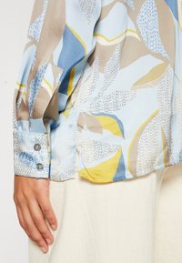 Blouse à manches longues en tissu bleu clair avec des motifs abstraits en beige, jaune et marine. Boutons décoratifs sur les poignets. Texture lisse.