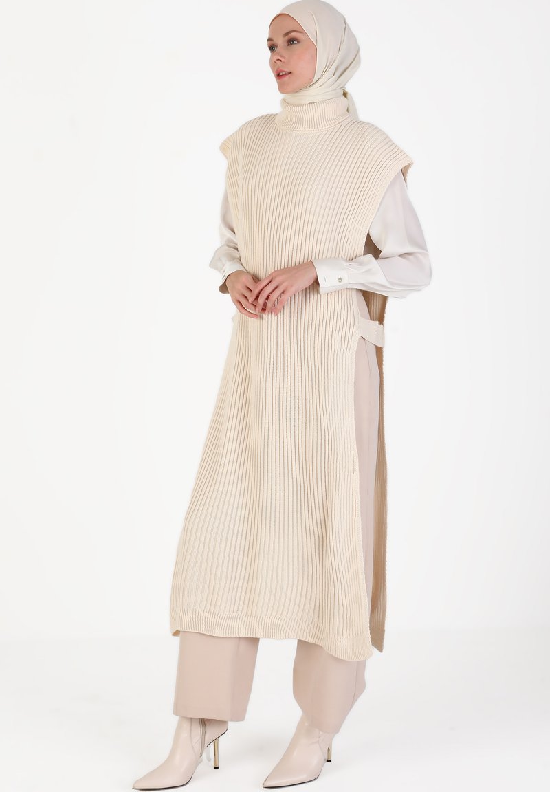 Beige ribbed knit tunik, jossa on korkea kaulus, ilman hihoja, sivuhalkiot, ja se on yhdistetty vaaleanbeigeihin housuihin sekä teräväkärkisiin nilkkasaappaisiin.