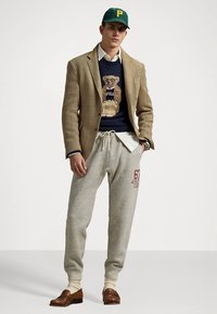 Μπλέιζερ με μοτίβο κουκίδας, μπλε πουλόβερ με εικόνα αρκούδας, γκρι φόρμα με κόκκινο κείμενο, καφέ μοκασίνια, πράσινο καπέλο μπέιζμπολ.