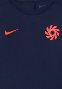 Marineblå sportskjorte med rød Nike-svask på venstre bryst og en rød stilisert sirkulær logo på høyre bryst.