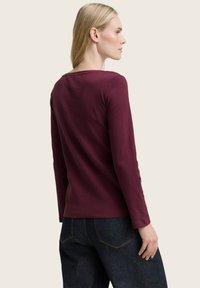 Frau mit blonden Haaren, die ein langärmeliges burgunderfarbens Oberteil und dunkelblaue Jeans trägt, von hinten vor einem einfarbigen Hintergrund gezeigt.