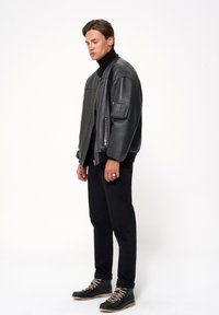 Czarna skórzana kurtka bombowiec z wysokim kołnierzem, widocznym zamkiem błyskawicznym i teksturowanymi łatami, zestawiona z czarnymi spodniami i sznurowanymi butami.