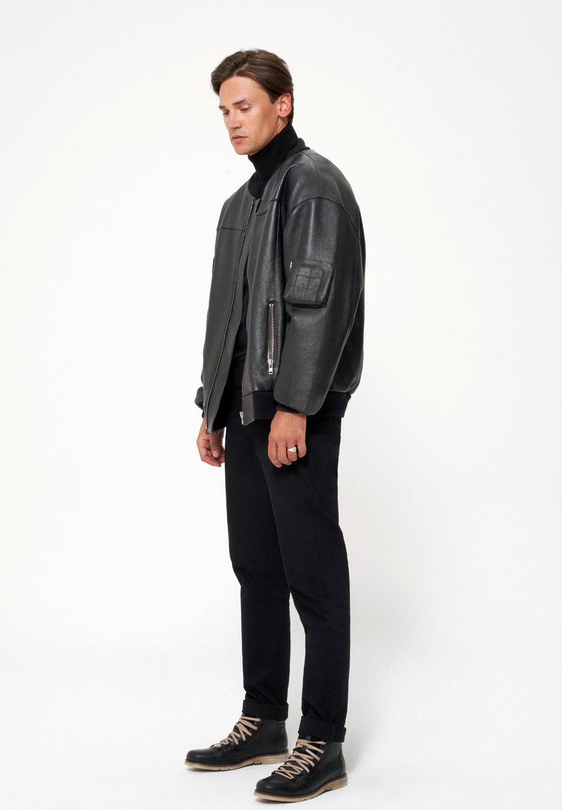 Czarna skórzana kurtka bombowiec z wysokim kołnierzem, widocznym zamkiem błyskawicznym i teksturowanymi łatami, zestawiona z czarnymi spodniami i sznurowanymi butami.