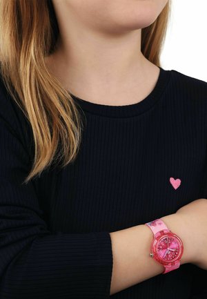 Girl with light brown hair wearing a black top with a small pink heart and a pink analog watch on her wrist.