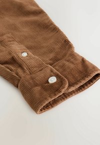 Manche en tissu velours côtelé marron avec boutons blancs au poignet et détail de patte de boutonnage sur fond clair.
