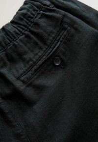 Primo piano di pantaloni in tessuto scuro con elastico in vita e tasca a filetto con bottone sul retro.