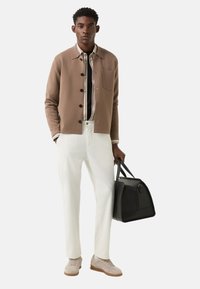 Hellbraune Strickjacke, schwarzes Hemd, weiße Hose; graue Sneaker und schwarze Ledertasche. Glatte Textur, Knopfdetails, lässiger Stil.