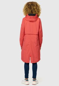 Chaqueta larga de color coral con capucha, cintura ajustable y abertura trasera, combinada con jeans de color azul oscuro y zapatillas blancas.