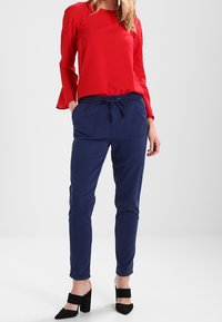 Červený top s dlouhým rukávem a rozšířenými manžetami, doplněný o tmavě modré jogger kalhoty s elastickým pasem a bočními kapsami, obutý s černými podpatky.