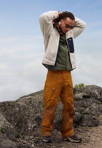 Minkštas baltas švarkas su juodu užtrauktuku kišene, alyvuogių žalios spalvos bazinė apranga ir oranžinės spalvos krovinių kelnės su kišenėmis bei rudomis kelių detalėmis. Juodi batai.