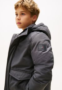 Giacca a vento grigia con cappuccio, caratterizzata da una finitura testurizzata, tasca anteriore e un patch con "Tommy Hilfiger" sulla manica.