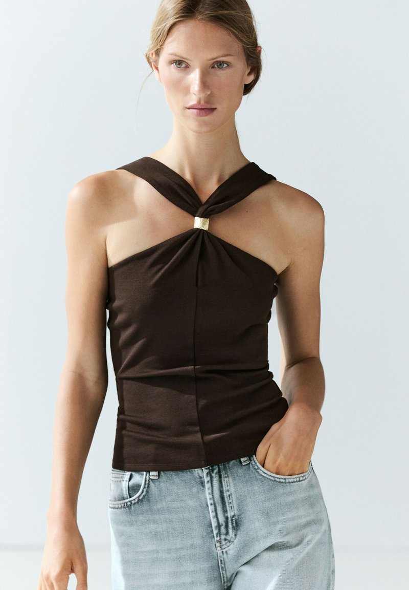 Woman wearing a sleeveless brown top with a gold clasp at the neckline and light blue denim jeans, standing against a plain background.