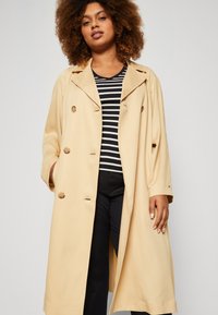 Trench beige de doble botonadura con grandes botones, solapas anchas y un ajuste relajado. Llevado sobre una camiseta de rayas en blanco y negro.