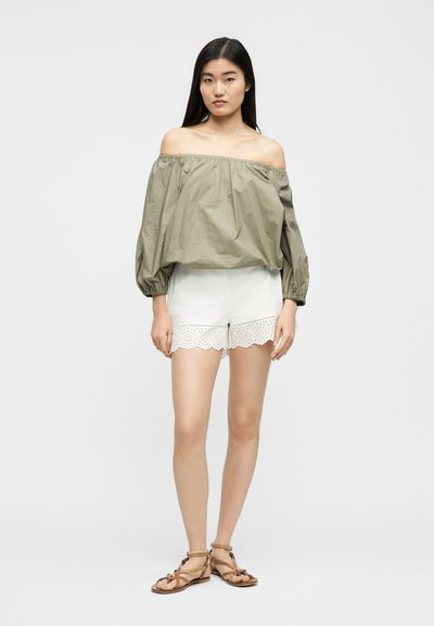 Γυναίκα στέκεται φορώντας ένα πράσινο τοπ χωρίς ώμους σε χρώμα ελιάς, λευκά σορτς από δαντέλα με τρύπες και ταμπά φλατ σανδάλια με λουριά σε ουδέτερο φόντο.