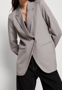 Vaaleanharmaa pikkutakki, jossa on istuva malli, yksi nappisuljenta ja kaksi etutaskua. Sujuva pinta ja rakenteelliset kaulukset.