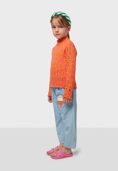 Camisa de manga larga naranja brillante con un estampado de girasoles, combinada con pantalones azul claro con un diseño gráfico y sandalias rosas.