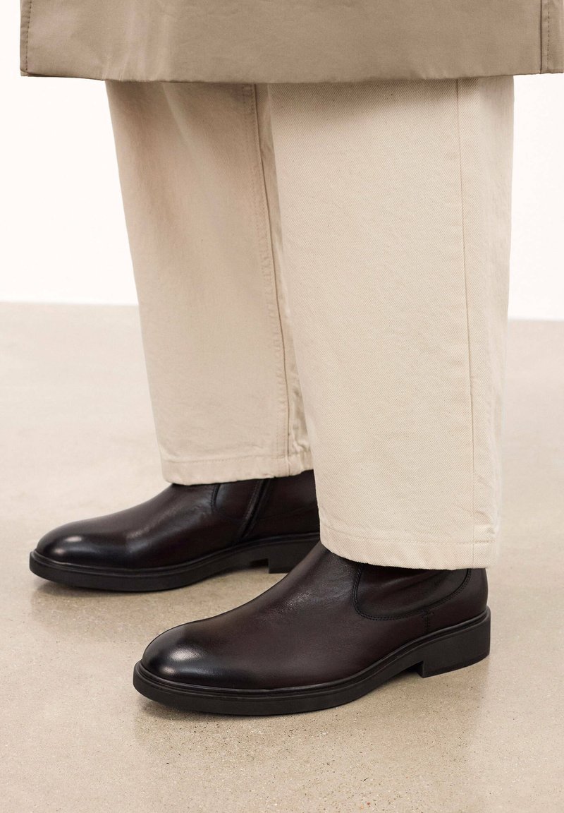 Botas de tobillo de cuero negro con un acabado suave, suela resistente y paneles laterales de elastano, combinadas con pantalones de pierna ancha en un tono beige claro.