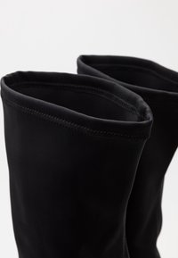 Une paire de bottes noires en tissu doux vue de dessus, mettant en avant la texture lisse et le point zigzag le long du bord supérieur.