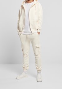 Felpa con cappuccio color crema con cerniera e pantaloni cargo abbinati, entrambi in un materiale morbido. I pantaloni hanno tasche laterali e polsini affusolati. Scarpe da ginnastica bianche.