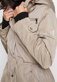 Beige prošívaná bunda s kapucí, se stříbrnými detaily, zipovou kapsou, elastickým pasem a logem na rukávu.