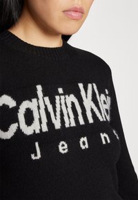Jeune personne portant un pull en maille noire avec un grand texte blanc "Calvin Klein Jeans" sur le devant, visible des épaules aux lèvres.