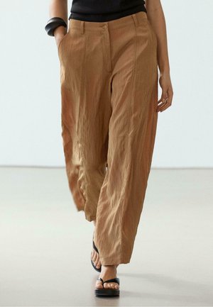 Person wearing loose brown pants, black sleeveless top, black bangle, and black platform sandals, walking on a light floor with hands relaxed.