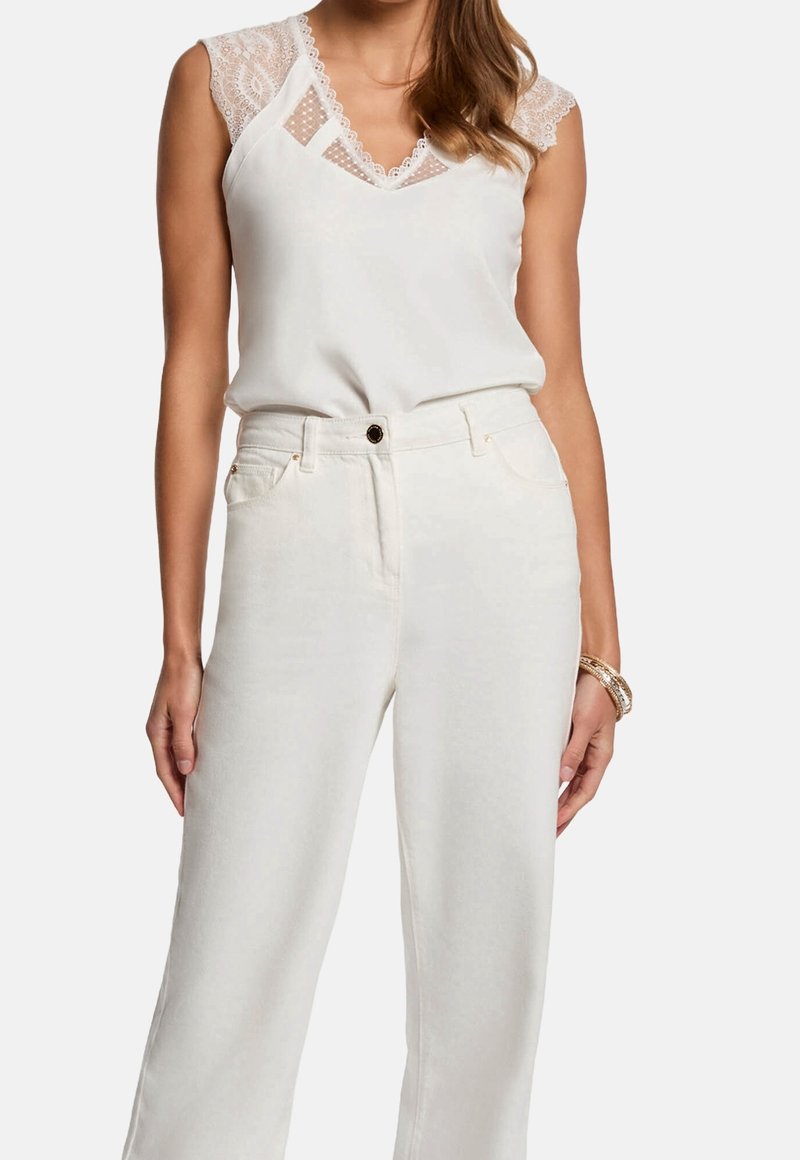 Femme portant un chemisier blanc sans manches orné de dentelle, rentré dans un pantalon blanc large à taille haute, avec des bracelets dorés au poignet.