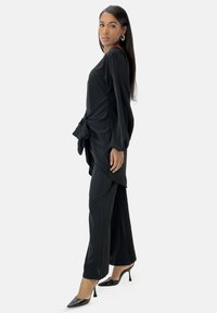 Tunika nera fluida con maniche lunghe e dettaglio con legatura laterale, abbinata a pantaloni larghi coordinati e scarpe con tacco alto nere.