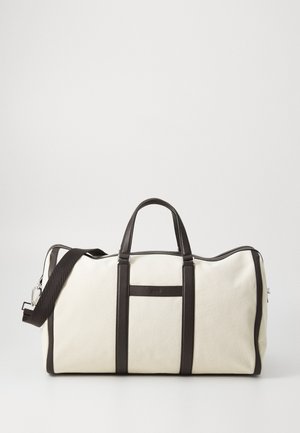 Canvas duffel bag in cream, featuring dark brown leather accents and handles, a front pocket, and a removable shoulder strap.