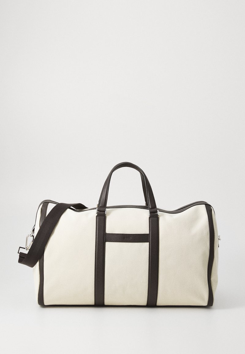 Borsa da viaggio in tela di colore crema, con dettagli e manici in pelle marrone scuro, tasca anteriore e tracolla rimovibile.