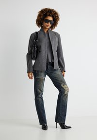 Γυναίκα που φοράει υπερμεγέθη σκούρο γκρι σακάκι, γκρι μπλούζα, σκισμένο τζιν με τρουκς, μαύρες μυτερές ψηλοτάκουνες, μεγάλα γυαλιά ηλίου και κρατάει μαύρη τσάντα ώμου.