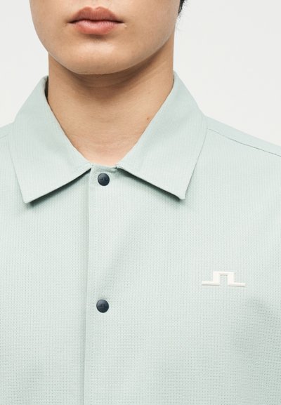 Κοντινό πλάνο ενός ατόμου που φοράει πουκάμισο ανοιχτού πράσινου χρώματος με υφή, γιακά, μαύρα κουμπιά και μικρό λευκό λογότυπο στο στήθος.